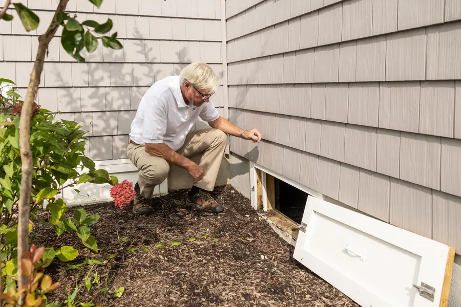 Inspector kneeling at an open crawl space vent beside a house